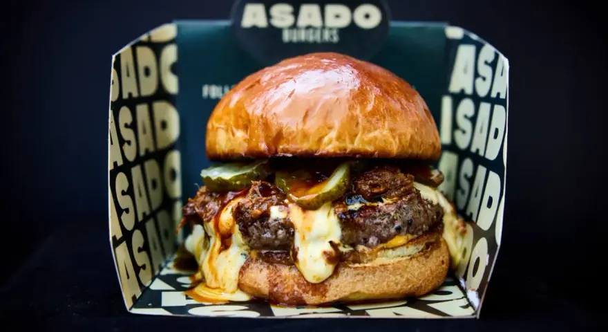 Saftiger Burger mit geschmolzenem Käse und Gurken, präsentiert in einer "ASADO BURGERS" Box. Der beste Burger Kölns wartet!