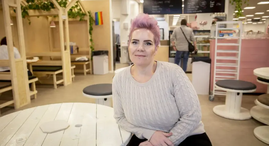 Woman with pink hair and a light sweater sits at a white wooden table in a cafe.