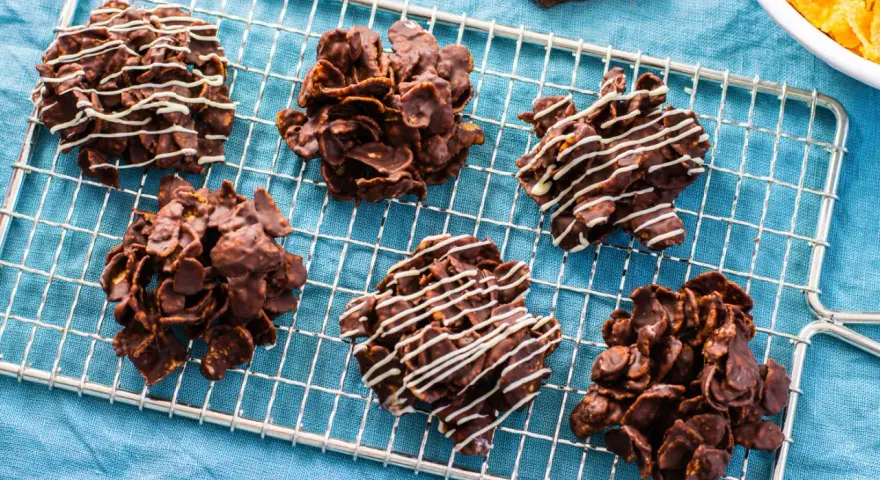 Sei dolcetti croccanti al cioccolato, simili a rose del deserto, raffreddano su una griglia. Alcuni sono decorati con sottili fili di cioccolato bianco.