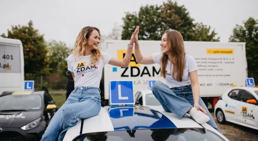 Dwie uśmiechnięte dziewczyny świętują sukces na dachu samochodu szkoleniowego. W tle ciężarówka z informacją o kursach na różne kategorie prawa jazdy.