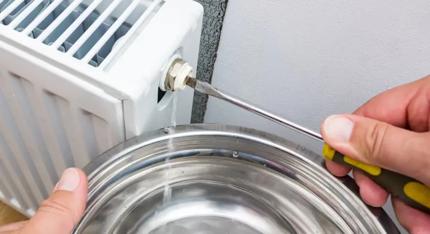 Osoba odkręca odpowietrznik grzejnika śrubokrętem, a woda kapie do metalowej miski. To pokazuje, jak odpowietrzyć grzejnik.