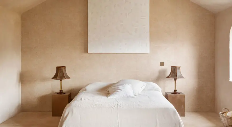 Camera da letto aesthetic con pareti color sabbia, letto bianco e due lampade in vimini. Un quadro astratto completa l'arredo.