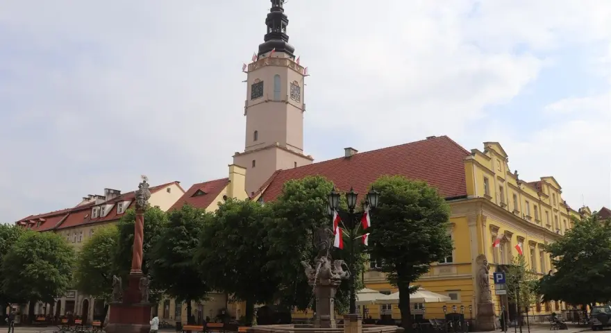 Wieża ratuszowa w Świdnicy góruje nad rynkiem, ozdobiona polskimi flagami. Obok fontanna i zabytkowe kamienice.