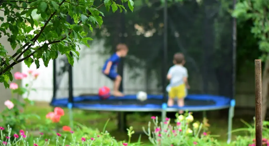 Dzieci bawią się na trampolinie w ogrodzie, otoczone zielenią i kwiatami.
