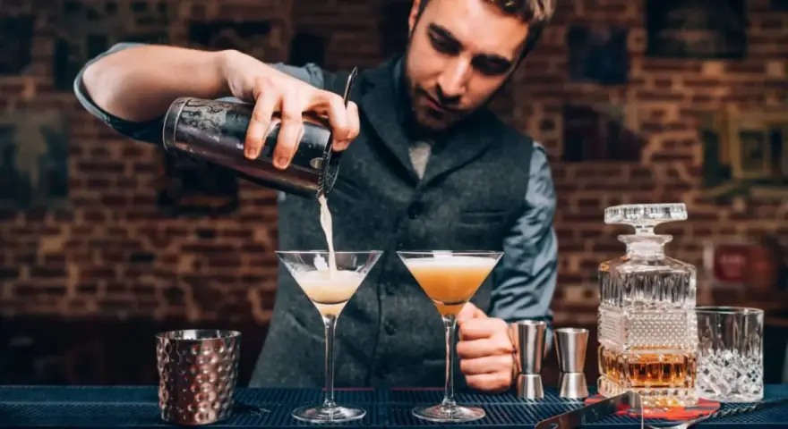 Barman przelewa drinka do kieliszka, pokazując, jak być dobrym barmanem. Na blacie stoją też inne drinki i akcesoria.