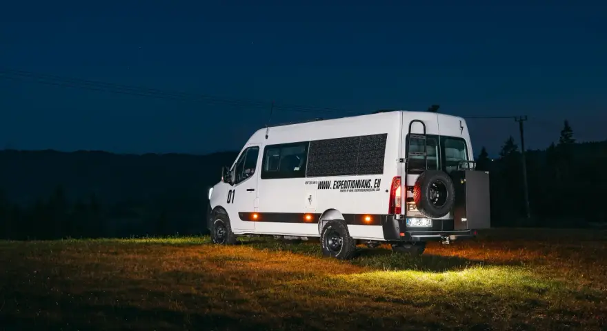 Biały kamper Renault Master z kołem zapasowym z tyłu, zaparkowany na łące nocą.