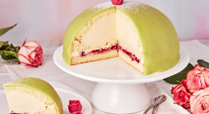 Ein Anschnitt eines grünen Prinzessinnenkuchens mit Sahnefüllung und Himbeermarmelade, verziert mit einer Zuckerrose. Ein Stück liegt auf einem Teller daneben.