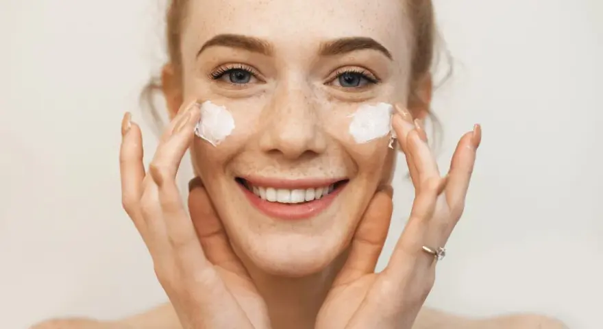 Uśmiechnięta dziewczyna z piegami nakłada krem na twarz. Sudocrem na co? Na suchą skórę, podrażnienia, zaczerwienienia.