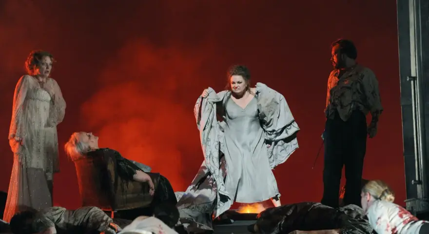 Scen från en grekisk teater med skådespelare i dramatiska poser. En kvinna i ljus klänning, en annan i grå klänning med tyg i famnen, och en man i mörk kostym.