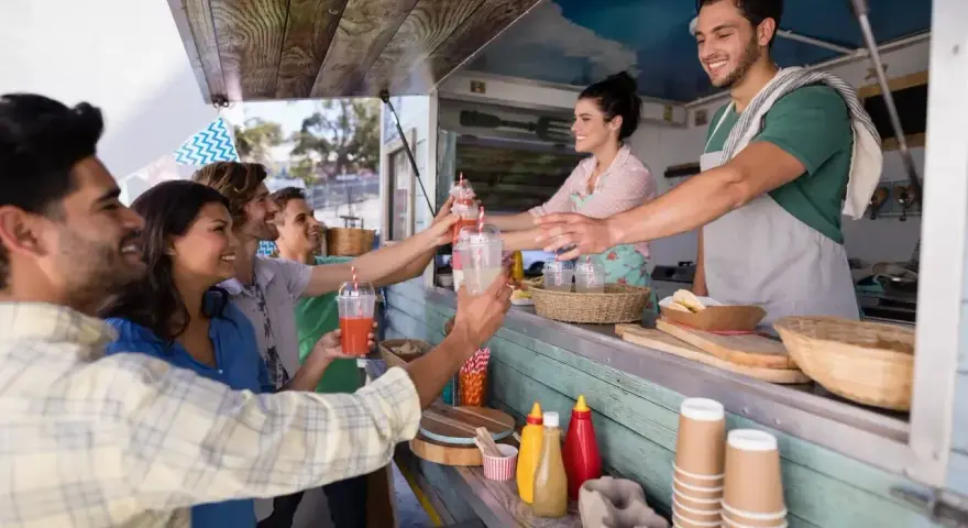 Uśmiechnięci klienci odbierają napoje od barmana w food trucku. Ciekawe, ile zarabia barman w takim miejscu?