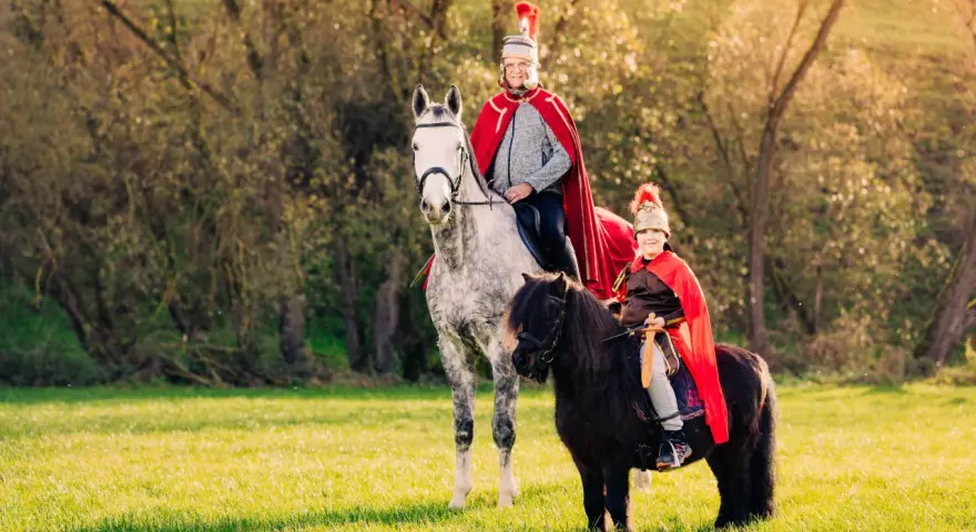 Zwei Reiter in römischer Kleidung, ein Erwachsener auf einem Schimmel und ein Kind auf einem Pony, reiten auf einer sonnigen Wiese. Ein fröhlicher Anblick, wie bei einem Martin Fest.