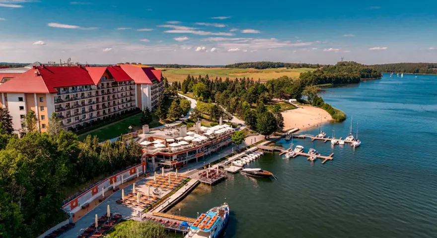Hotel na wyspie Mikołajki z czerwonym dachem, otoczony zielenią, z mariną pełną łodzi i statkiem wycieczkowym na jeziorze.