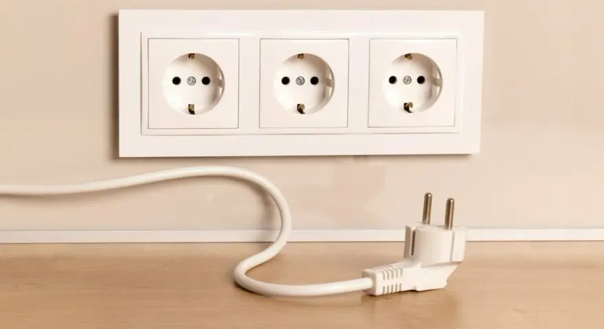 Biały kabel z wtyczką leży na drewnianym blacie obok potrójnego gniazdka. Jak schować kable, by wyglądały estetycznie?