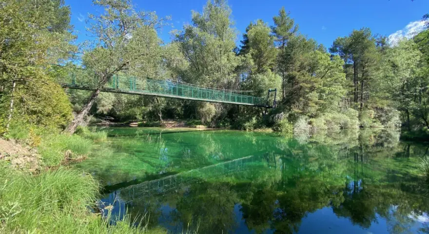 Puente verde cruza un río de aguas cristalinas y verdes, reflejando el bosque exuberante. Un lugar idílico para explorar el **alto tajo**.