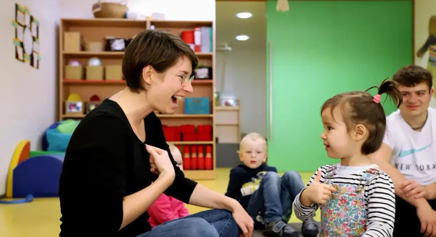 Eine Frau lacht und gestikuliert, während sie mit einem Kleinkind sprechen übt. Ein Junge und ein Mann sitzen im Hintergrund.