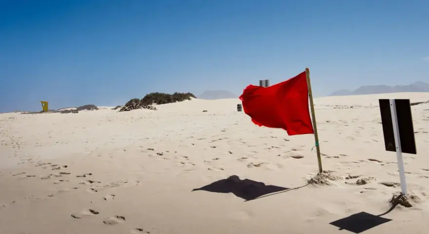 Czerwone flagi na plaży ostrzegają przed niebezpieczeństwem. Wiatr rozwiewa materiał, tworząc dynamiczny obraz na tle błękitnego nieba i piaszczystego krajobrazu.