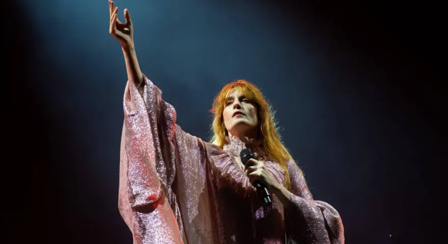 Florence Welch z Florence and the Machine na koncercie, w błyszczącej sukni, z uniesioną ręką.