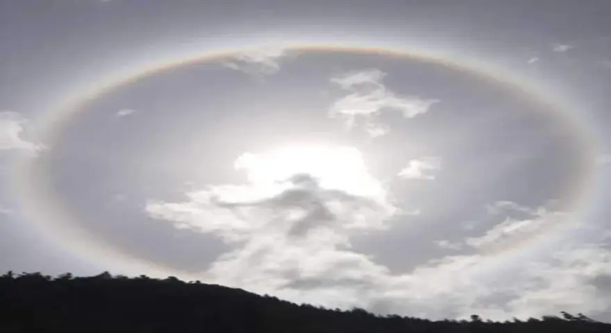 Un alone arcobaleno circonda il sole tra le nuvole, creando un effetto magico sopra una collina scura.