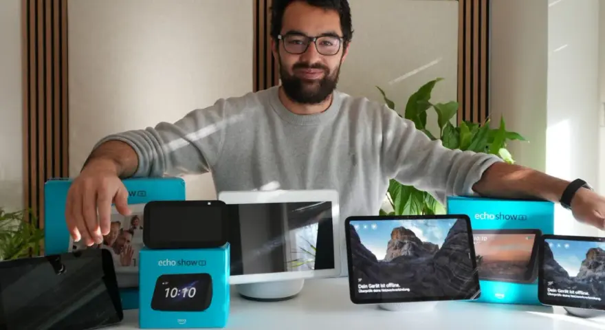 Mann präsentiert verschiedene Echo Show Generationen. Mehrere Geräte und Verpackungen sind auf dem Tisch zu sehen.
