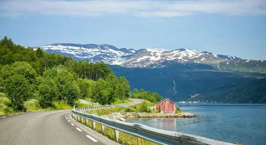 En slingrande väg längs en fjord i Norge, med snöklädda berg i bakgrunden. En röd stuga vid vattnet. Perfekt för en roadtrip norge rutt.