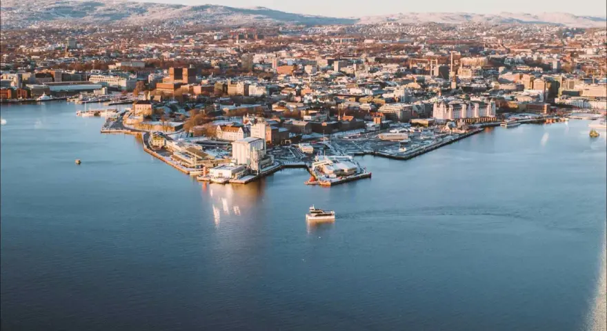 Widok na zimowe Oslo z wody. Mała łódka płynie po spokojnym akwenie, w tle rozciąga się miasto i ośnieżone wzgórza.