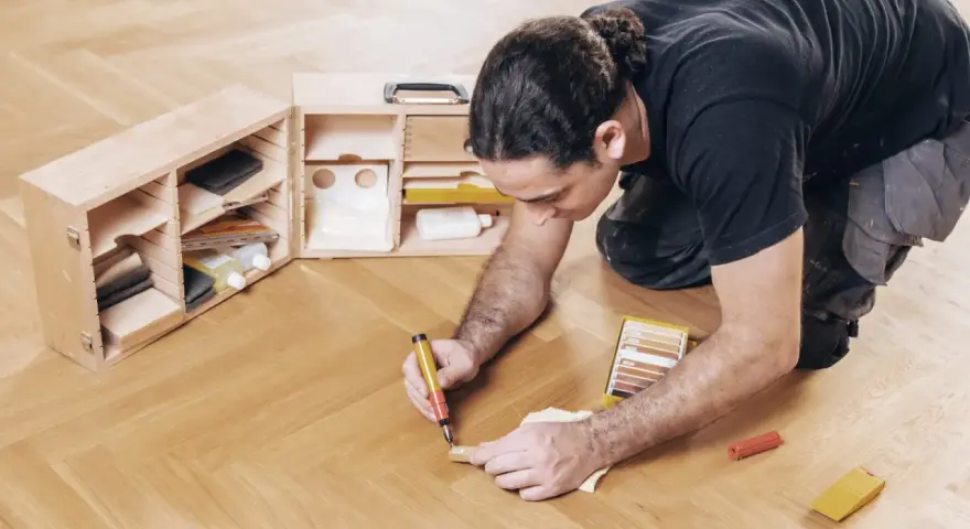 Mann repariert Parkett mit einem Reparaturstift und einer Farbauswahl. Werkzeugkasten mit Zubehör daneben.