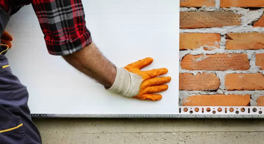 Ręka w pomarańczowej rękawicy przykłada płytę pir do ceglanej ściany.