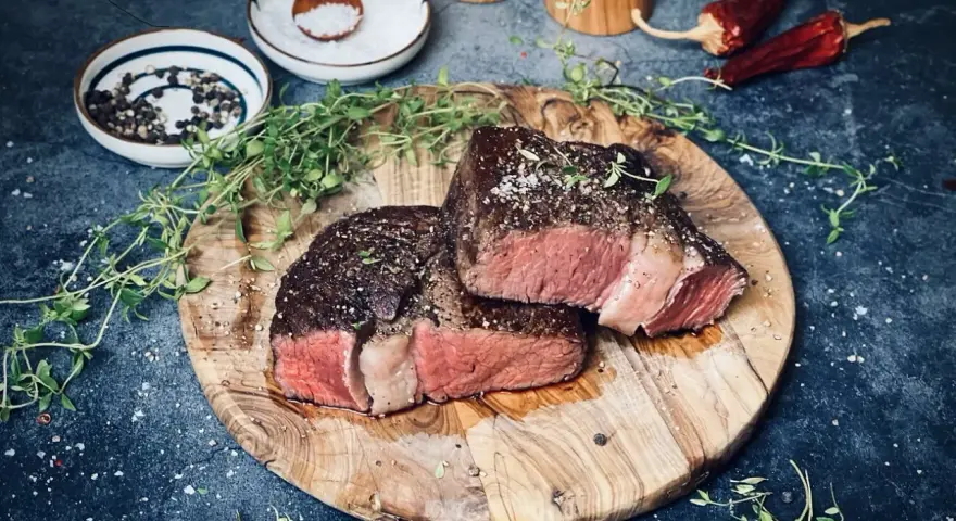 Zwei saftige Rib-Eye-Steaks, perfekt gegrillt, liegen auf einem Holzbrett. Mit Meersalz und Pfeffer bestreut, garniert mit frischem Thymian.