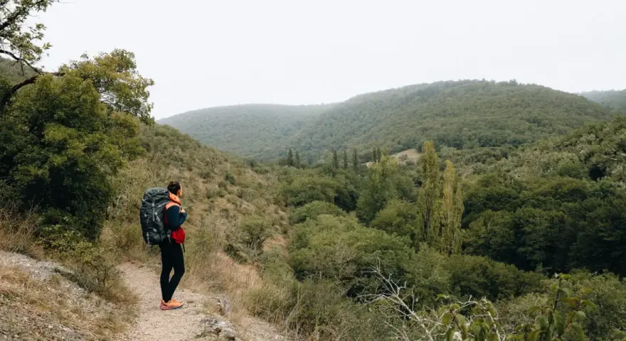 Randonneuse avec sac à dos sur un chemin d'amadou, contemplant une vallée verdoyante. La carte du chemin d'amadou est dans sa poche.