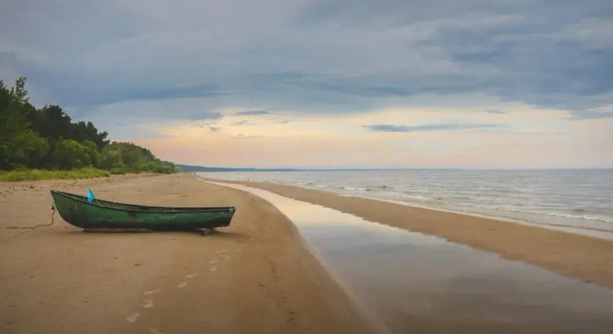 Zielona łódź na piaszczystej plaży Mierzei Wiślanej, idealne miejsce na camping. Woda odbija niebo, tworząc spokojny krajobraz.