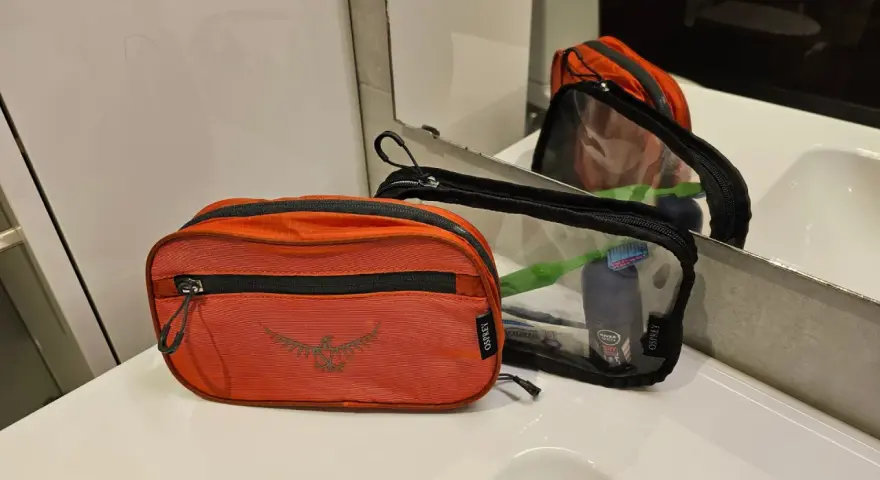 Två Osprey-necessärer, en orange och en genomskinlig, ligger på en vit bänkskiva. Den genomskinliga necessären innehåller tandborste och deodorant.
