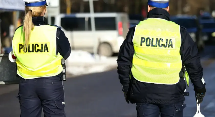 Dwóch policjantów w kamizelkach odblaskowych z napisem "POLICJA" stoi na drodze.