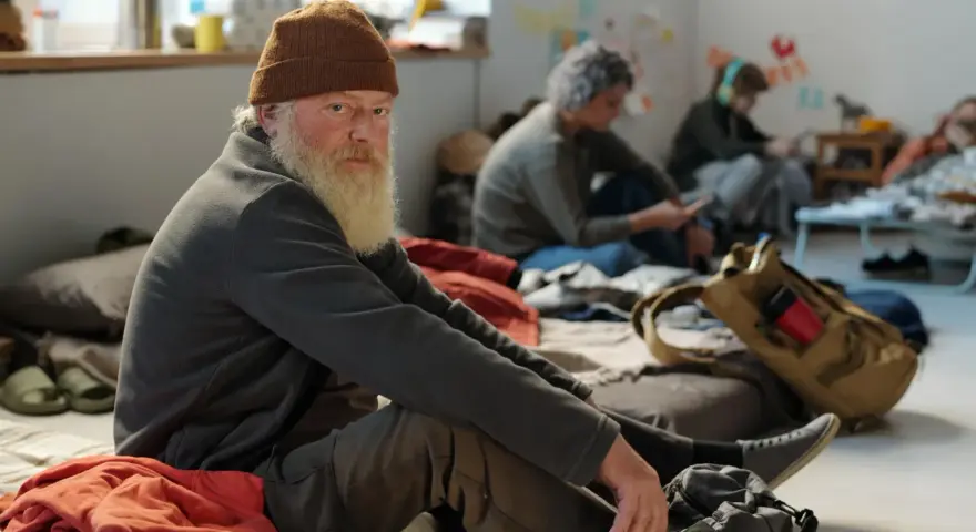 Älterer Mann mit Bart und Mütze sitzt auf dem Boden, umgeben von anderen Menschen. Eine Szene, die die Frage aufwirft: Wohin ohne Geld und Wohnung?
