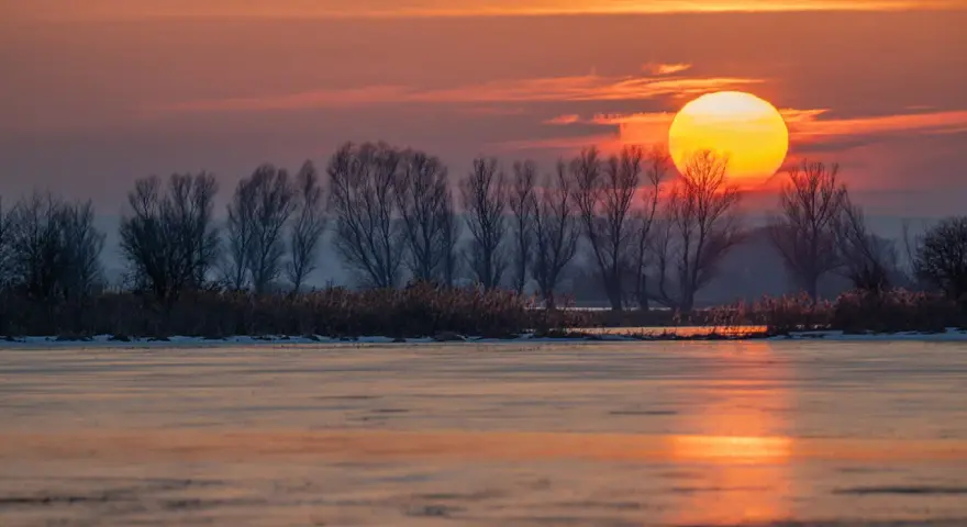 Zachód słońca nad zamarzniętym jeziorem. Piękna fotografia krajobrazowa z pomarańczowym niebem i sylwetkami drzew.