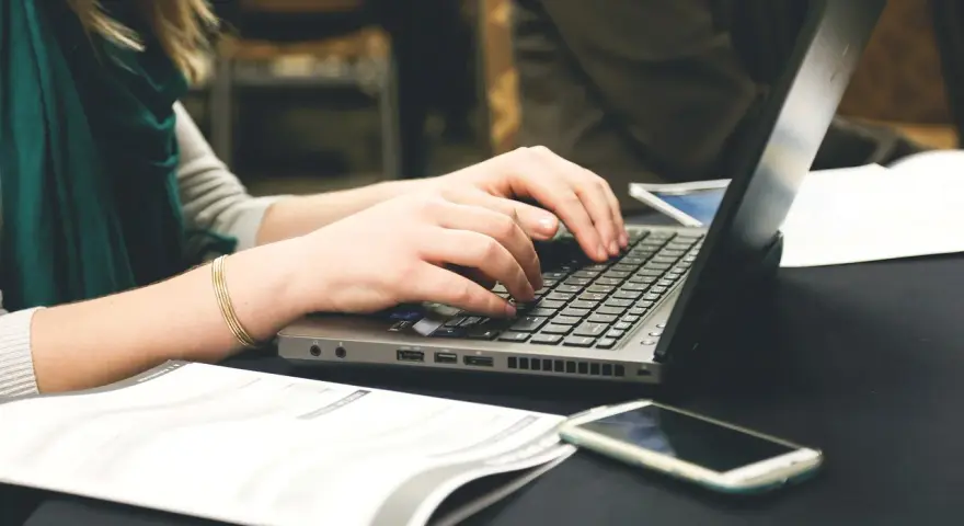 Dłonie piszą na laptopie, obok leży telefon i dokumenty. Może to być praca nad statutem szkoły.