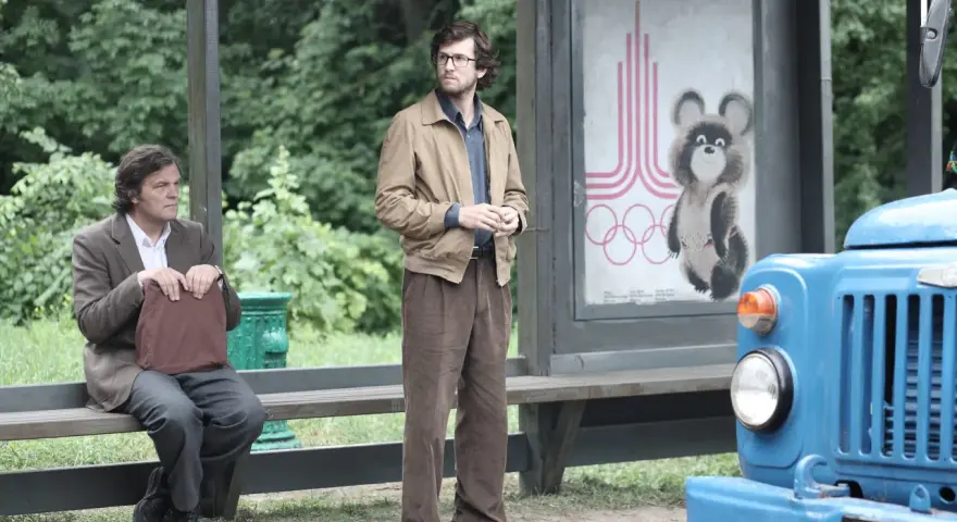 Deux hommes attendent à un arrêt de bus, un panneau des JO de Moscou 1980 avec Misha l'ours en arrière-plan. L'affaire Farewell se déroule.