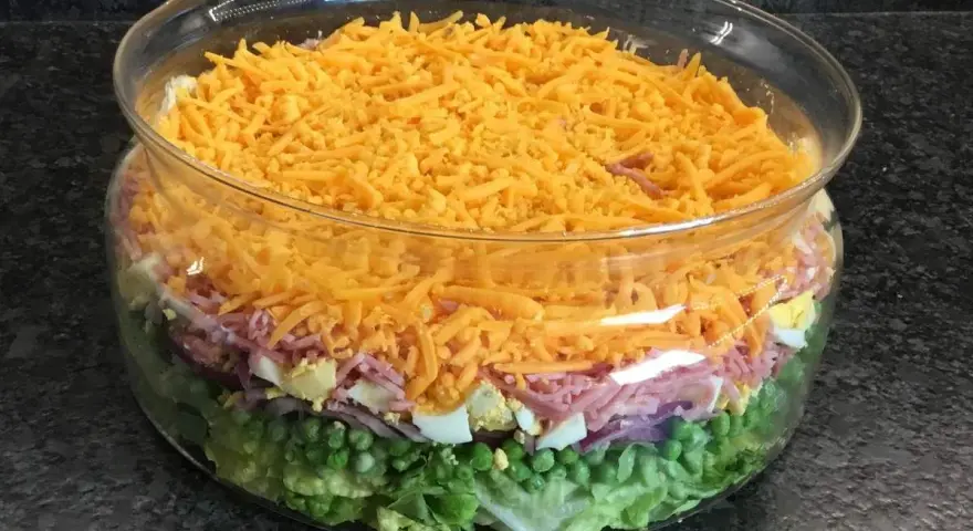 Une salade 7 étages colorée dans un grand bol en verre, avec des couches de laitue, petits pois, oignons rouges, jambon, œufs durs et fromage râpé.