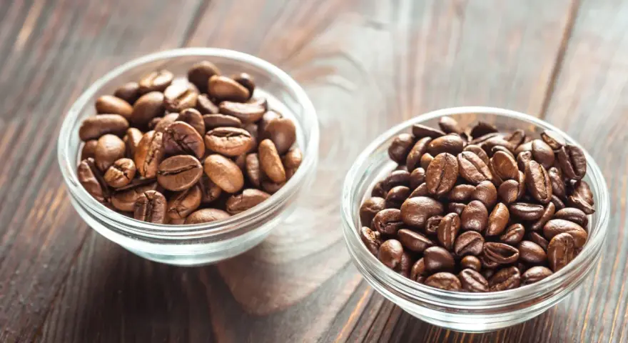 Dwie miseczki z ziarnami kawy: jaśniejsza arabika i ciemniejsza robusta, na drewnianym tle.