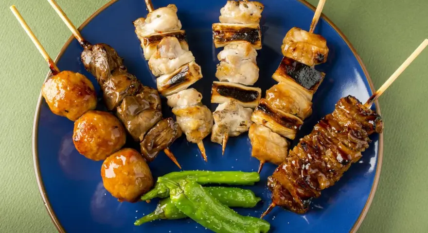 Różnorodne yakitori na niebieskim talerzu: kurczak, wątróbka, klopsiki i papryczki shishito.
