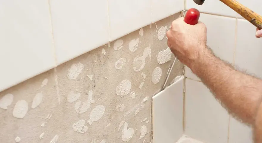 Main d'un bricoleur en pleine action pour enlever carrelage mural avec un burin et un marteau. La surface est prête pour un nouveau revêtement.
