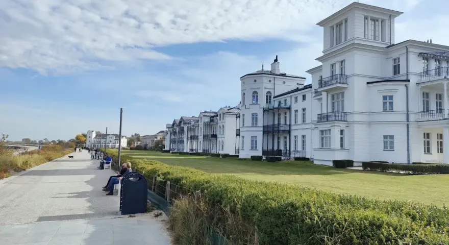 Weiße Villen säumen die Heiligendamm Promenade. Menschen spazieren und genießen die Aussicht auf das Meer.