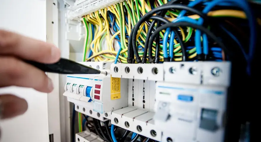 Ręka majstruje przy bezpiecznikach. Kolorowe przewody tworzą skomplikowaną instalację elektryczną.