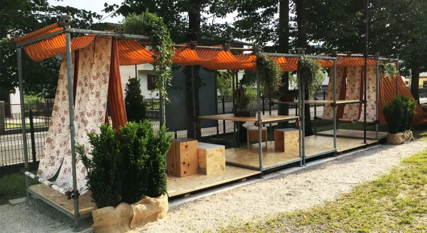 Struttura esterna con tende a righe e floreali, tavoli e sedute in legno. Un esempio di come costruire un gazebo con tubi innocenti, pratico e decorativo.