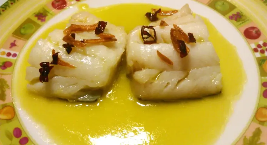Dos trozos de bacalao al pil pil, cubiertos con ajo frito y guindilla, nadan en una salsa dorada sobre un plato decorado.
