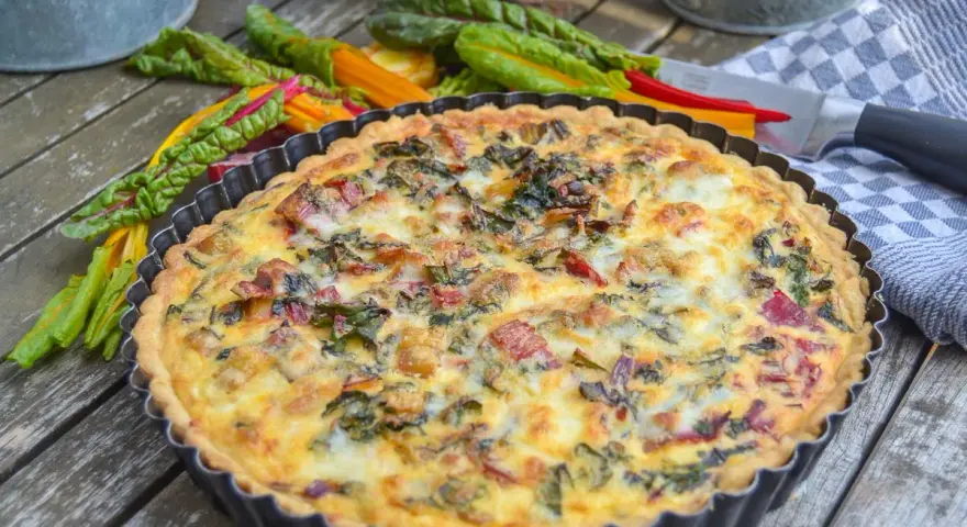 Herzhaftes Mangold Quiche Rezept, frisch gebacken und bereit zum Genießen. Bunte Mangoldstiele liegen daneben.