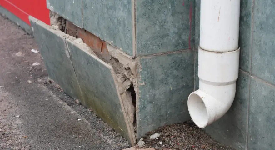 Beschädigte Sockelabdichtung am Haus. Fliesen lösen sich, darunter Ziegelmauerwerk. Ein Fallrohr leitet Wasser ab.