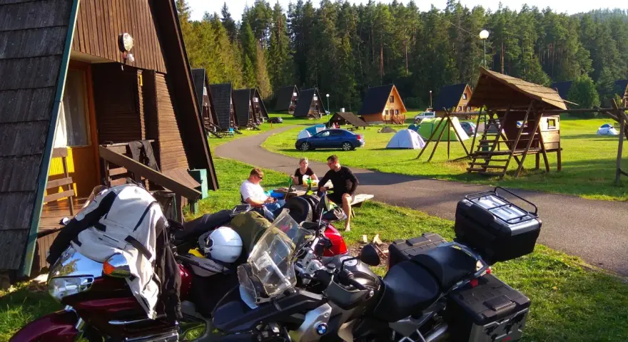 Motocykliści odpoczywają na kempingu w Rumunii, otoczeni drewnianymi domkami i lasem.