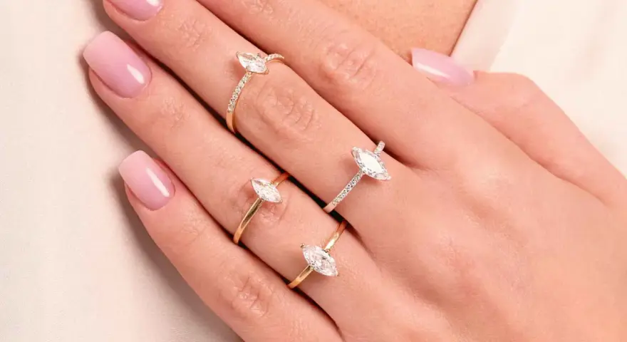 Dłonie z różowym manicure ozdobione są trzema złotymi pierścionkami z białymi kamieniami. Jeden z nich to pierścionek kwadratowy.