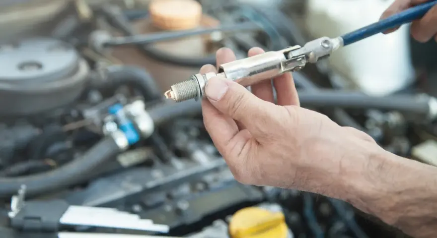 A hand holds a spark plug with a socket wrench attached, ready for a spark plug replacement. This DIY task can save on the spark plug replacement cost.
