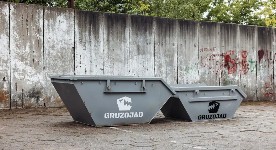 Dwa szare kontenery na śmieci zmieszane, z logo GRUZOJAD, stoją przed zniszczonym betonowym murem pokrytym graffiti.